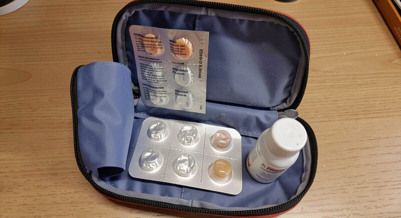 An open first aid pouch with blister packs, a bandage roll and a small bottle arranged on a table.